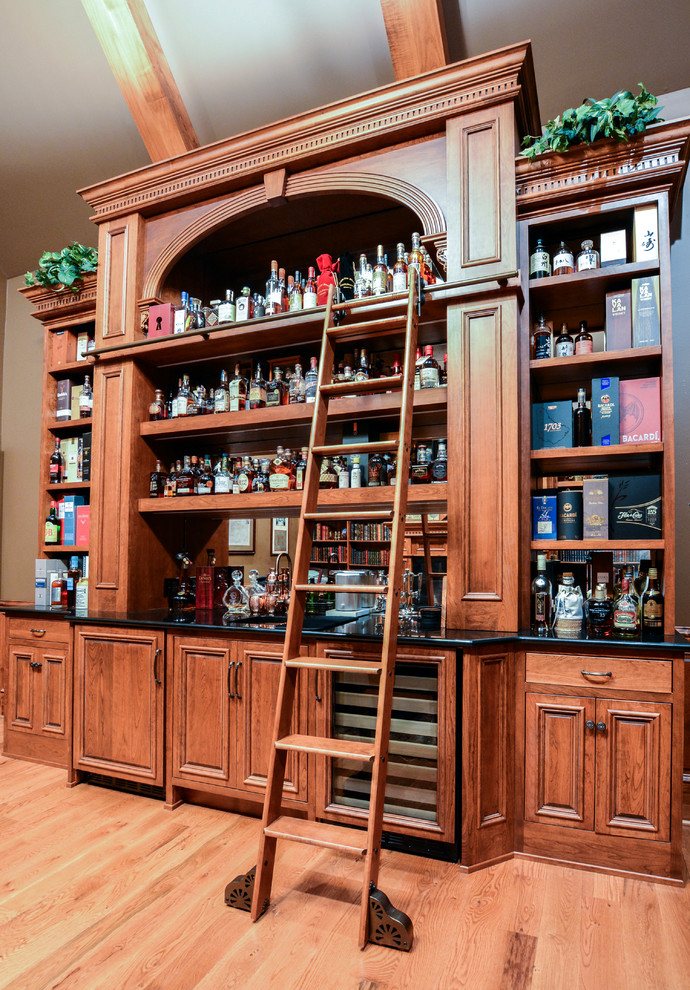 Photo of a mid-sized traditional galley home bar in Other with raised-panel cabinets, brown cabinets, granite benchtops, mirror splashback and light hardwood floors.