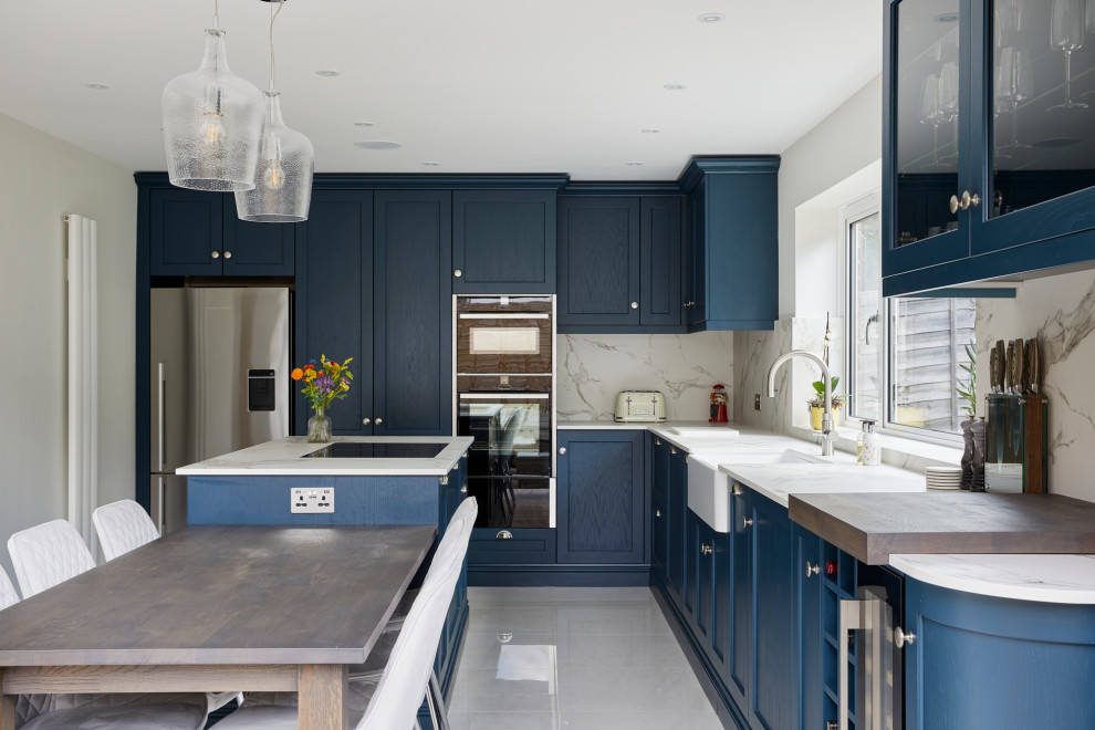 Design ideas for a large contemporary l-shaped kitchen/diner in London with a belfast sink, recessed-panel cabinets, blue cabinets, engineered stone countertops, white splashback, stone slab splashback, stainless steel appliances, an island and white worktops.