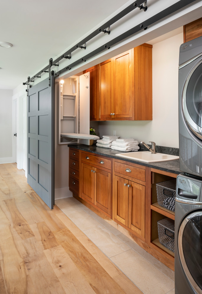 Inspiration for a mid-sized transitional single-wall dedicated laundry room in Detroit with a drop-in sink, shaker cabinets, medium wood cabinets, laminate benchtops, white walls, light hardwood floors, a stacked washer and dryer, beige floor and grey benchtop.