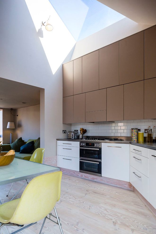 Small contemporary eat-in kitchen in London with flat-panel cabinets, white cabinets, stainless steel benchtops, white splashback, subway tile splashback and light hardwood floors.