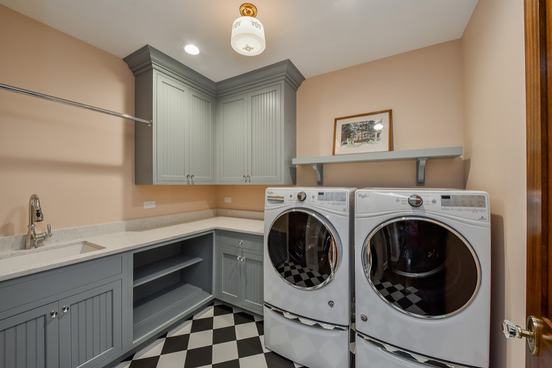 Inspiration for a small traditional l-shaped dedicated laundry room in Chicago with an undermount sink, beaded inset cabinets, blue cabinets, quartz benchtops, pink walls, ceramic floors, a side-by-side washer and dryer, black floor and white benchtop.