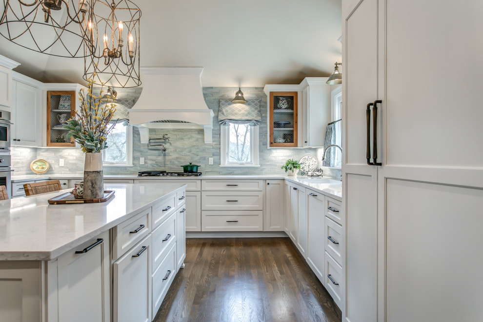 Foto på ett stort vit kök, med skåp i shakerstil, vita skåp, blått stänkskydd, mellanmörkt trägolv, en köksö och brunt golv