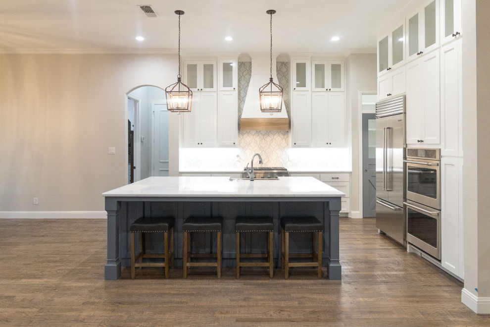 Inspiration for a large transitional l-shaped medium tone wood floor and brown floor open concept kitchen remodel in Dallas with a farmhouse sink, shaker cabinets, white cabinets, quartz countertops, white backsplash, mosaic tile backsplash, stainless steel appliances, an island and white countertops
