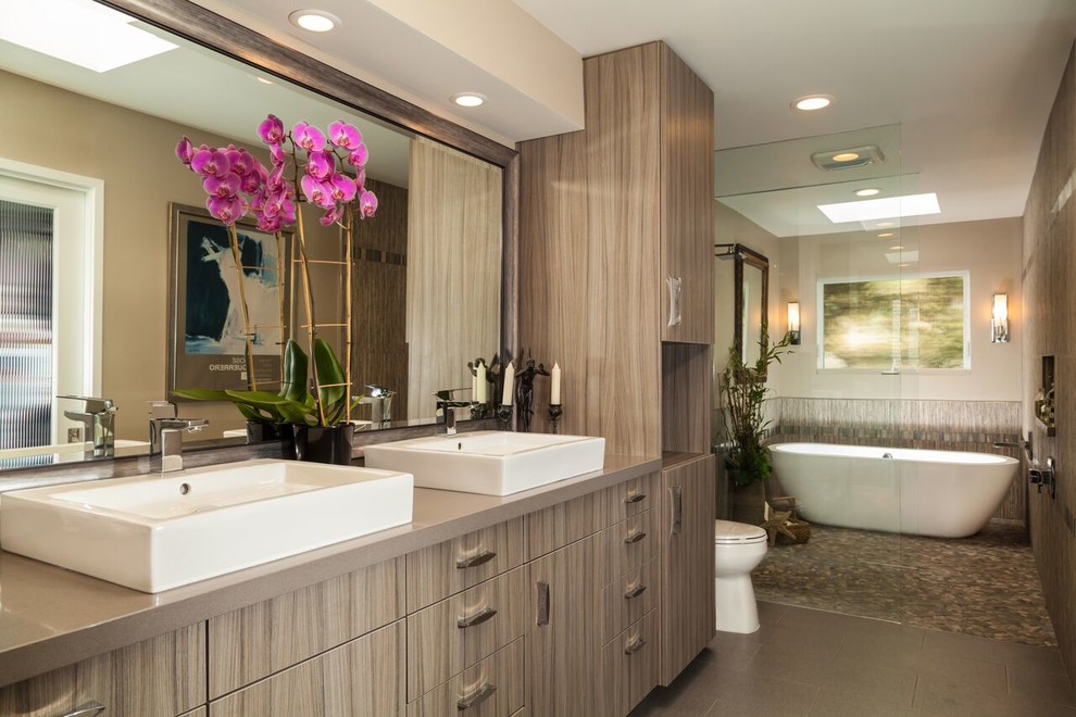 Photo of a mid-sized contemporary master bathroom in Los Angeles with flat-panel cabinets, light wood cabinets, engineered quartz benchtops, a trough sink, a freestanding tub, a curbless shower, a one-piece toilet, gray tile, mosaic tile, grey walls and pebble tile floors.