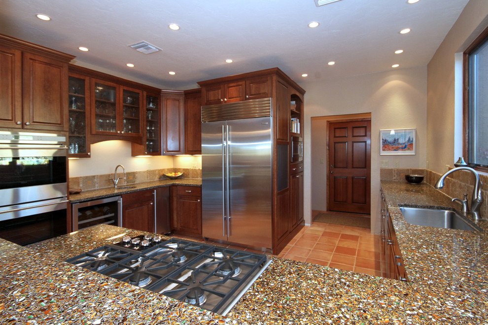 Photo of a large transitional u-shaped eat-in kitchen in Phoenix with a single-bowl sink, raised-panel cabinets, brown cabinets, recycled glass benchtops, beige splashback, stone tile splashback, stainless steel appliances and terra-cotta floors.
