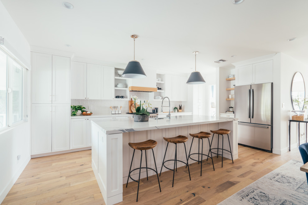 Photo of a country l-shaped open plan kitchen in Los Angeles with a farmhouse sink, shaker cabinets, white cabinets, white splashback, stainless steel appliances, medium hardwood floors, with island, brown floor and grey benchtop.