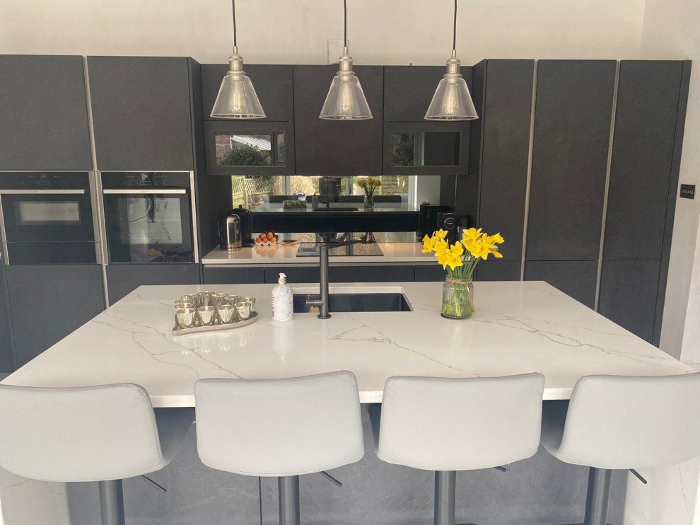 Large contemporary single-wall open plan kitchen in Manchester with a drop-in sink, flat-panel cabinets, grey cabinets, quartzite benchtops, grey splashback, mirror splashback, black appliances, ceramic floors, with island, grey floor and white benchtop.