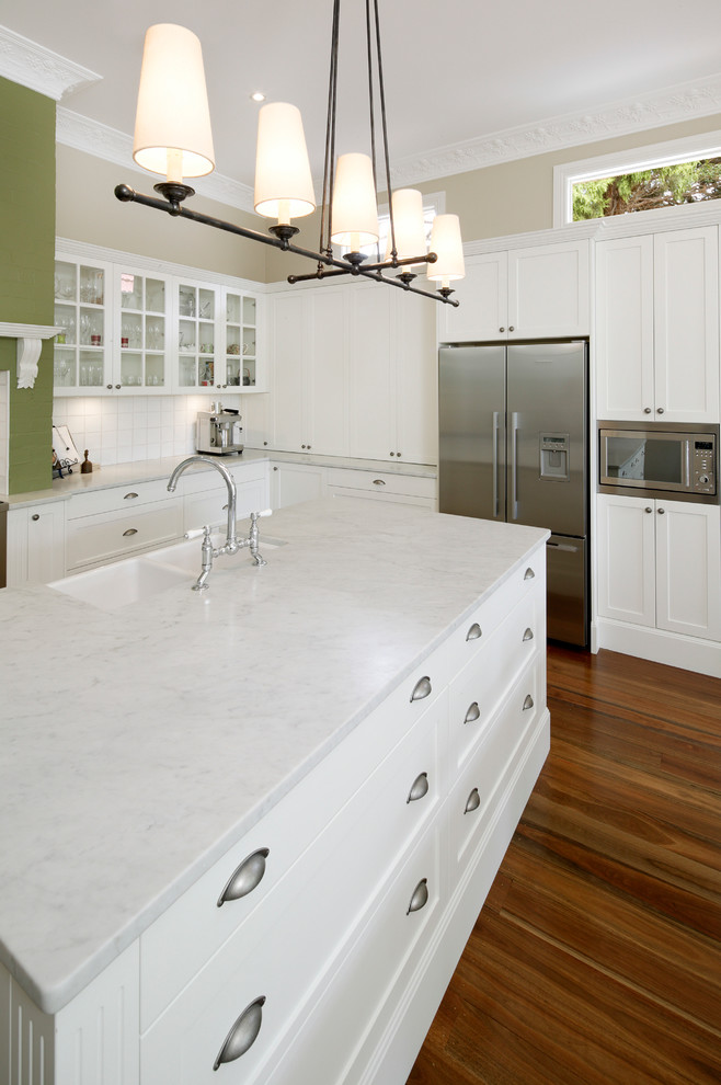 Design ideas for a large traditional l-shaped eat-in kitchen in Sydney with shaker cabinets, white cabinets, marble benchtops, white splashback, ceramic splashback, stainless steel appliances, with island, a farmhouse sink, medium hardwood floors, brown floor and white benchtop.