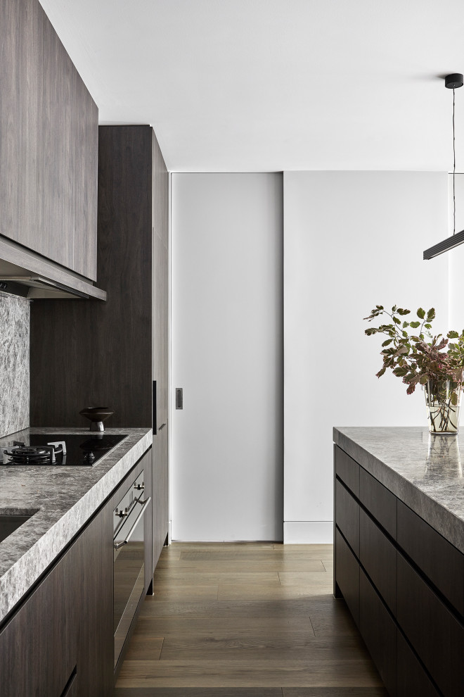 This is an example of a mid-sized modern open plan kitchen in Melbourne with an undermount sink, dark wood cabinets, limestone benchtops, grey splashback, limestone splashback, black appliances, light hardwood floors, with island, beige floor, grey benchtop and flat-panel cabinets.