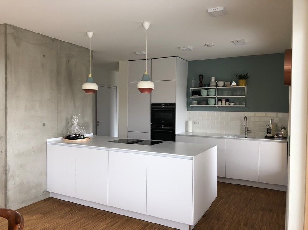 Zweizeilige, Mittelgroße, Offene Skandinavische Küche mit integriertem Waschbecken, weißen Schränken, Mineralwerkstoff-Arbeitsplatte, Rückwand aus Metrofliesen, schwarzen Elektrogeräten, braunem Holzboden, braunem Boden, weißer Arbeitsplatte, flächenbündigen Schrankfronten, Küchenrückwand in Weiß und Halbinsel in München