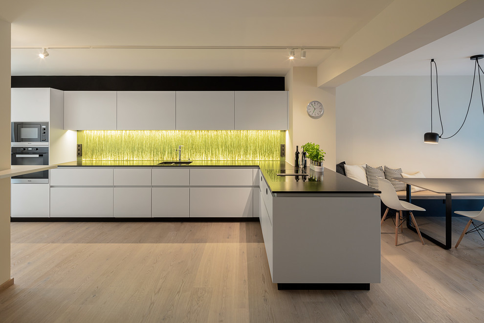 Photo of a large modern l-shaped open plan kitchen in Munich with flat-panel cabinets, white cabinets, a peninsula, an undermount sink, solid surface benchtops, green splashback, glass sheet splashback, black appliances, painted wood floors and brown floor.