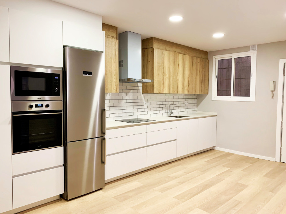 Foto på ett nordiskt vit linjärt kök med öppen planlösning, med en undermonterad diskho, släta luckor, skåp i mellenmörkt trä, vitt stänkskydd, stänkskydd i porslinskakel, rostfria vitvaror och mellanmörkt trägolv