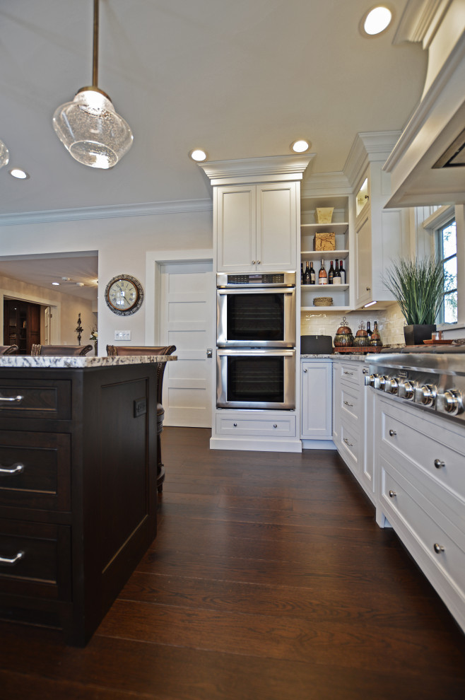 Example of a transitional dark wood floor and brown floor eat-in kitchen design with an undermount sink, shaker cabinets, white cabinets, granite countertops, white backsplash, ceramic backsplash, stainless steel appliances, an island and beige countertops