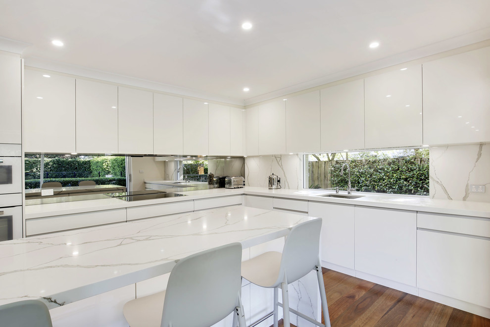 This is an example of a mid-sized modern l-shaped eat-in kitchen in Sydney with an undermount sink, flat-panel cabinets, white cabinets, quartz benchtops, metallic splashback, mirror splashback, white appliances, medium hardwood floors and with island.