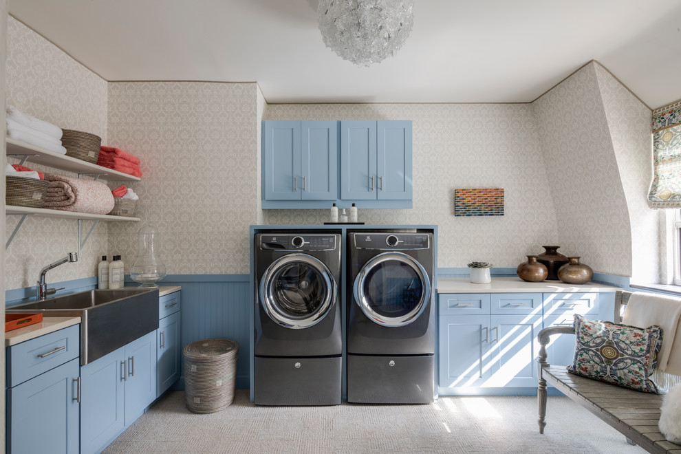 This is an example of a transitional l-shaped dedicated laundry room in New York with a farmhouse sink, shaker cabinets, blue cabinets, beige walls, carpet, a side-by-side washer and dryer, beige floor and beige benchtop.