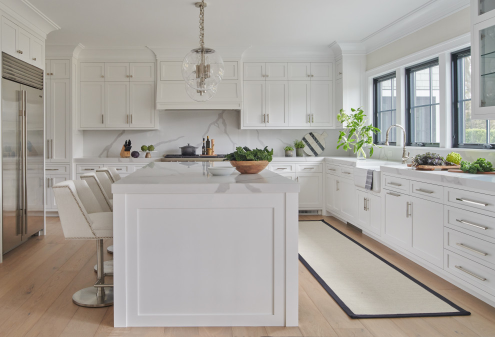 Inspiration for a transitional u-shaped kitchen in Other with a farmhouse sink, shaker cabinets, white cabinets, white splashback, stone slab splashback, stainless steel appliances, light hardwood floors, with island, beige floor and white benchtop.