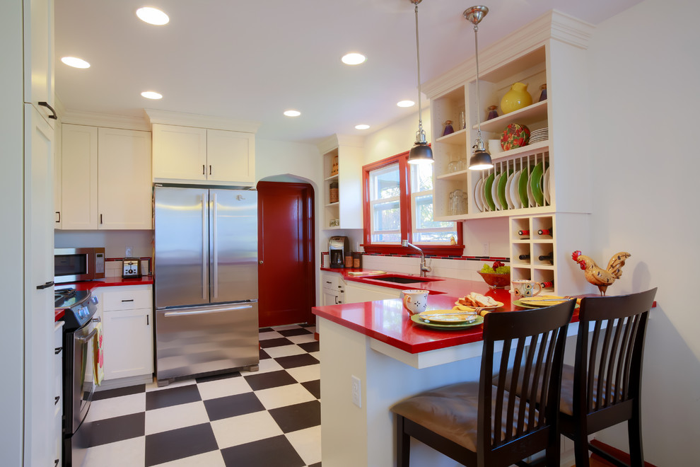 This is an example of a small eclectic l-shaped eat-in kitchen in Portland with an undermount sink, white splashback, subway tile splashback, stainless steel appliances, shaker cabinets, white cabinets, solid surface benchtops, a peninsula, black floor, red benchtop and vinyl floors.