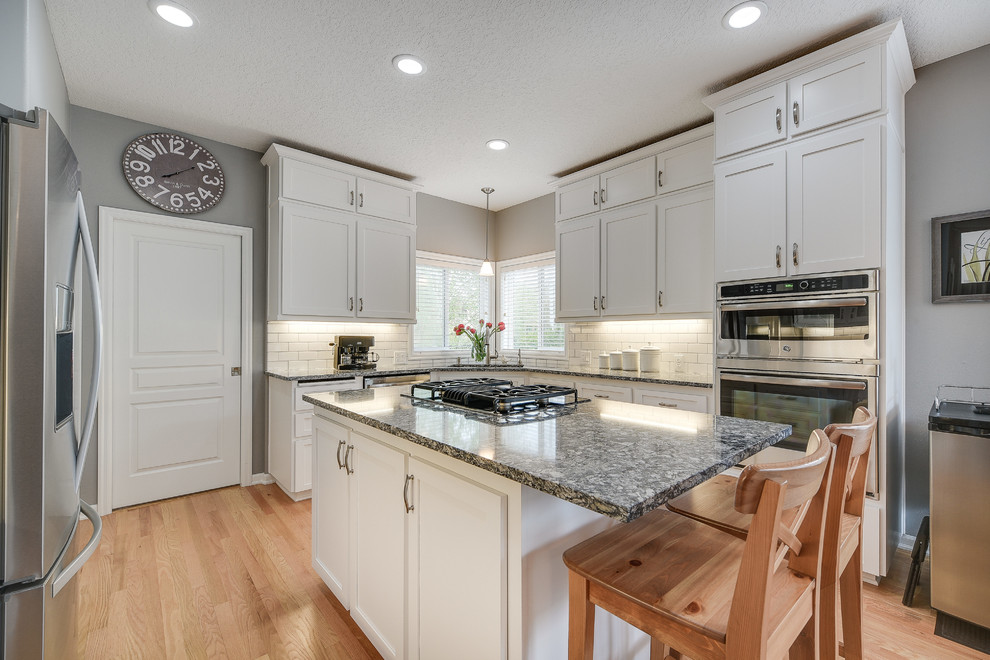 Example of a mid-sized transitional l-shaped light wood floor and brown floor enclosed kitchen design in Portland with a drop-in sink, shaker cabinets, white cabinets, quartz countertops, white backsplash, subway tile backsplash, an island and stainless steel appliances
