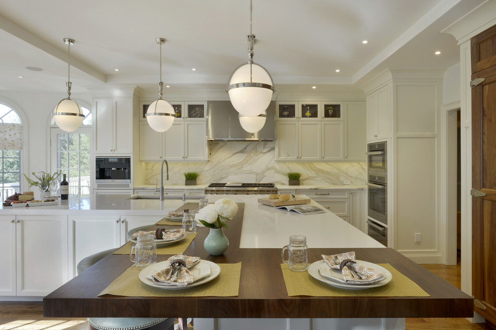 Large transitional l-shaped open plan kitchen in New York with an undermount sink, shaker cabinets, white cabinets, wood benchtops, white splashback, marble splashback, panelled appliances, medium hardwood floors, with island and brown floor.