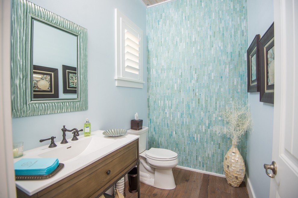 Inspiration for a mid-sized beach style powder room in Jacksonville with furniture-like cabinets, medium wood cabinets, a two-piece toilet, blue tile, green tile, glass tile, blue walls, medium hardwood floors, an integrated sink, solid surface benchtops and brown floor.