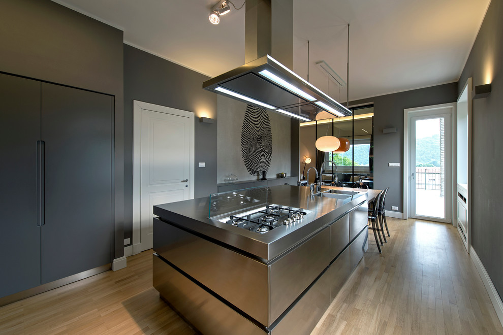 Inspiration for a contemporary separate kitchen in Milan with an integrated sink, flat-panel cabinets, stainless steel cabinets, stainless steel benchtops, stainless steel appliances, medium hardwood floors, with island and glass sheet splashback.