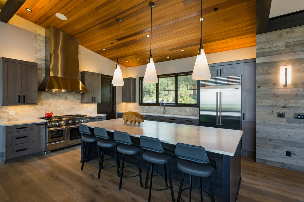 Large mountain style dark wood floor and brown floor kitchen photo in Albuquerque with shaker cabinets, quartzite countertops, beige backsplash, travertine backsplash, stainless steel appliances, an island, brown cabinets and beige countertops