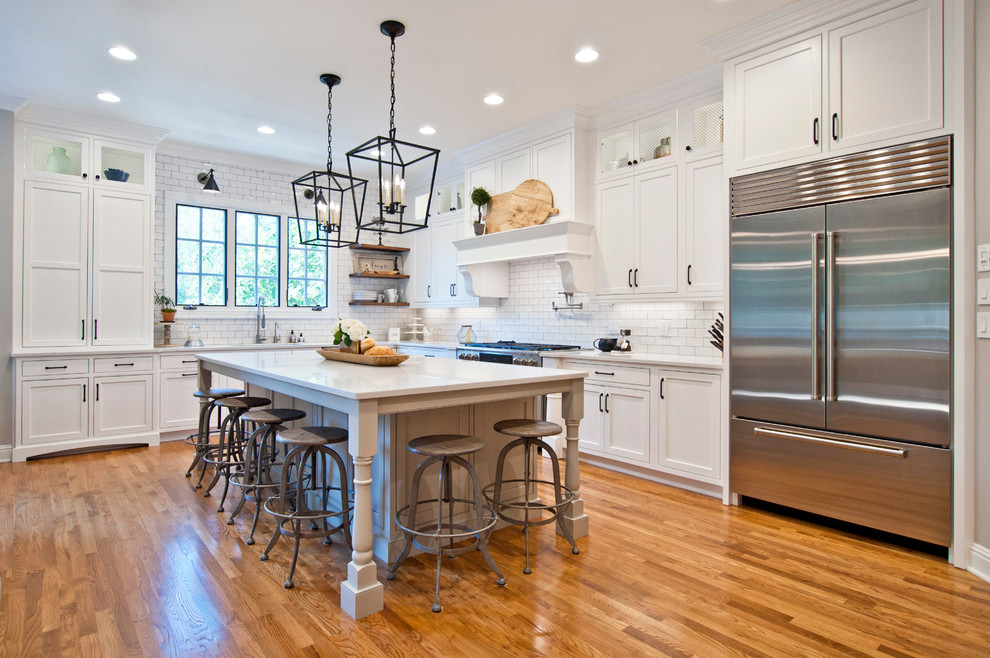 Design ideas for a large country l-shaped kitchen in Nashville with white cabinets, quartz benchtops, white splashback, subway tile splashback, stainless steel appliances, medium hardwood floors, with island, brown floor, white benchtop and shaker cabinets.