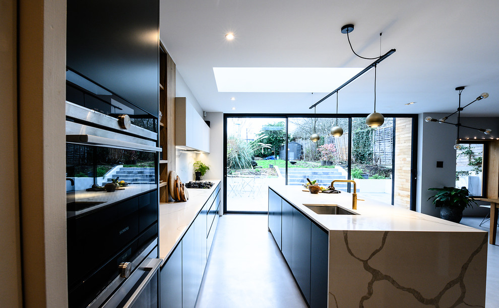 Inspiration for a large contemporary galley eat-in kitchen in London with flat-panel cabinets, green cabinets, white splashback, porcelain splashback, black appliances, concrete floors, with island and white benchtop.