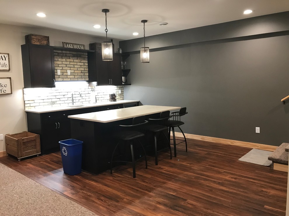 Inspiration for a country single-wall seated home bar in Milwaukee with an undermount sink, shaker cabinets, dark wood cabinets, quartz benchtops, beige splashback, brick splashback, vinyl floors, brown floor and beige benchtop.