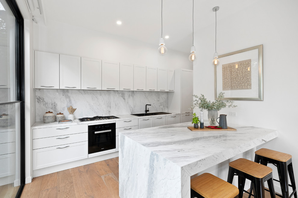 Inspiration for a mid-sized transitional single-wall open plan kitchen in Melbourne with a drop-in sink, white cabinets, marble benchtops, grey splashback, marble splashback, with island, grey benchtop, black appliances, medium hardwood floors and brown floor.