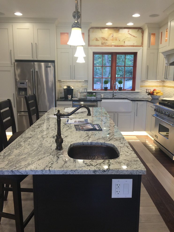 Example of a mid-sized transitional u-shaped light wood floor and beige floor eat-in kitchen design in Boston with recessed-panel cabinets, white cabinets, stainless steel appliances, an island, a farmhouse sink, granite countertops, beige backsplash, stone tile backsplash and multicolored countertops