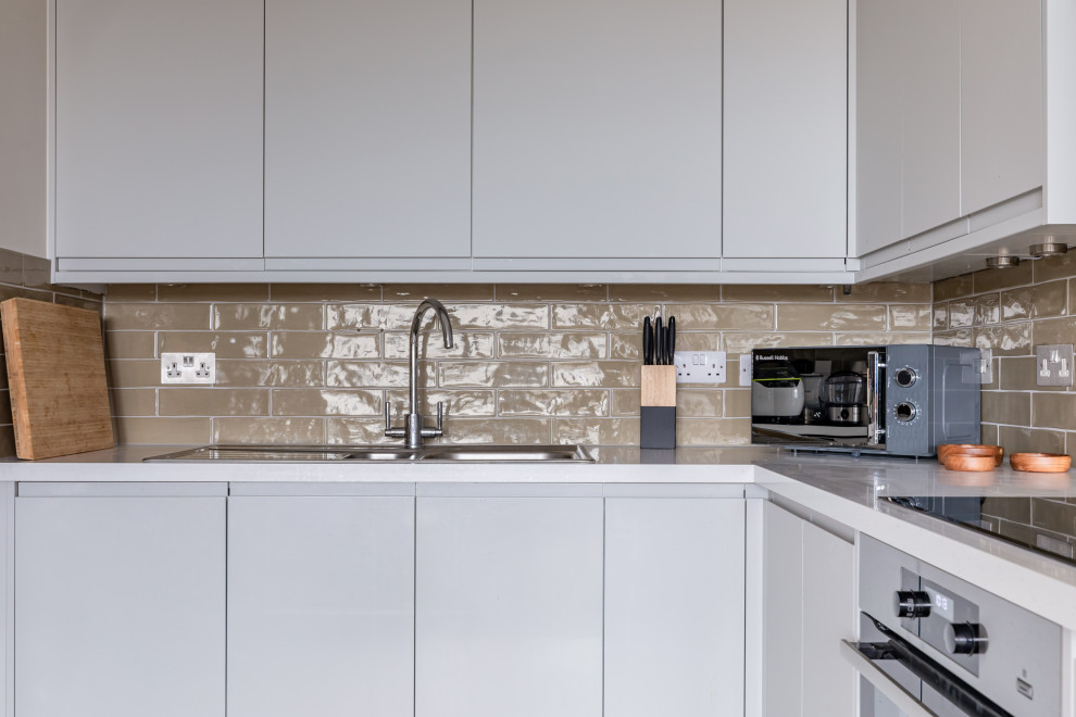 This is an example of a contemporary l-shaped open plan kitchen in London with flat-panel cabinets, grey cabinets, composite countertops, green splashback, ceramic splashback, integrated appliances and white worktops.