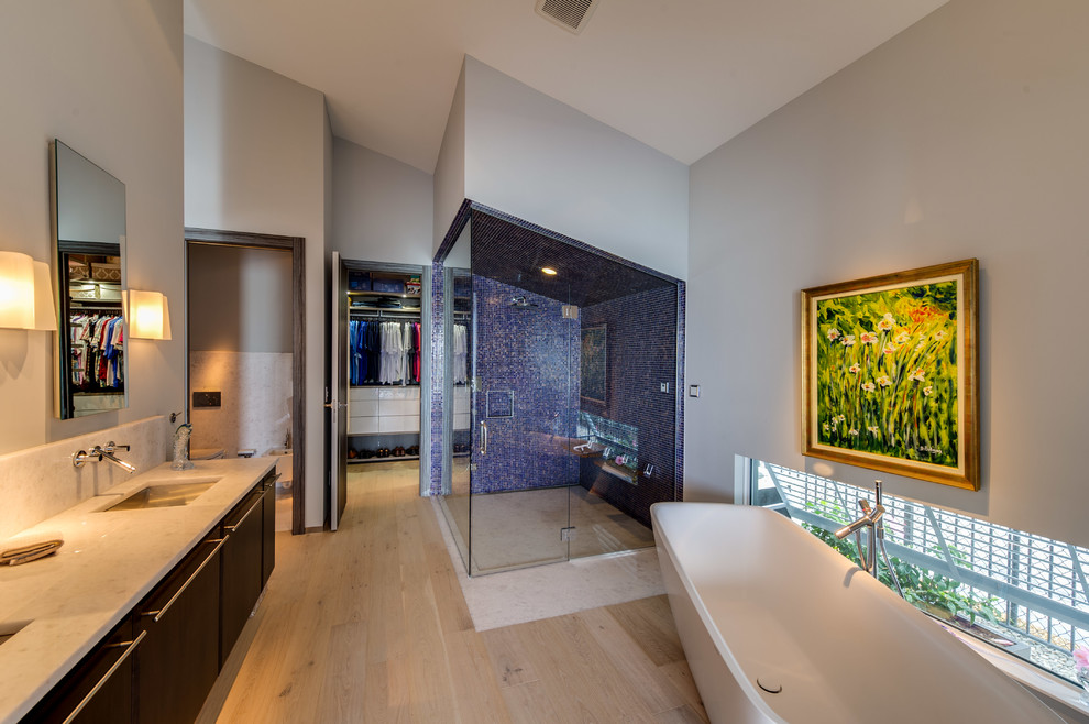 Expansive contemporary bathroom in Other with flat-panel cabinets, brown cabinets, a freestanding tub, a curbless shower, a wall-mount toilet, blue tile, glass tile, grey walls, light hardwood floors, an undermount sink and granite benchtops.