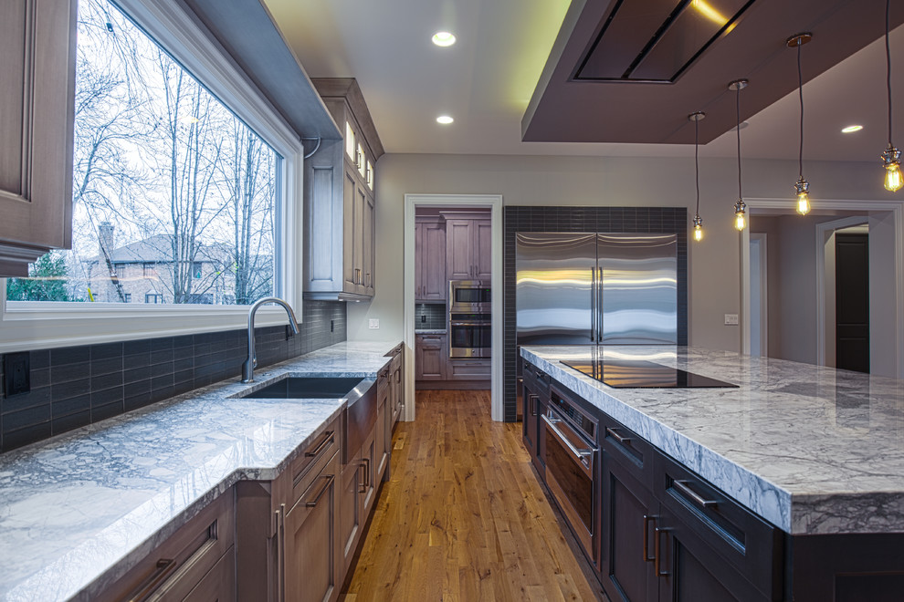 Photo of a large transitional l-shaped open plan kitchen in Detroit with recessed-panel cabinets, grey cabinets, stainless steel appliances, medium hardwood floors, with island, quartzite benchtops, grey splashback, glass tile splashback and a farmhouse sink.