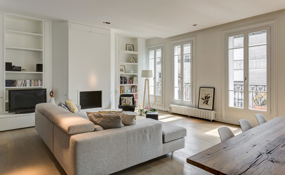 This is an example of a large scandinavian open concept family room in Paris with white walls, light hardwood floors, a standard fireplace, a freestanding tv, beige floor and a library.