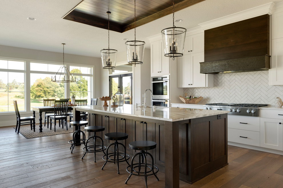 This is an example of a country eat-in kitchen in Minneapolis with an undermount sink, shaker cabinets, white cabinets, white splashback, stainless steel appliances, dark hardwood floors, with island, brown floor and white benchtop.