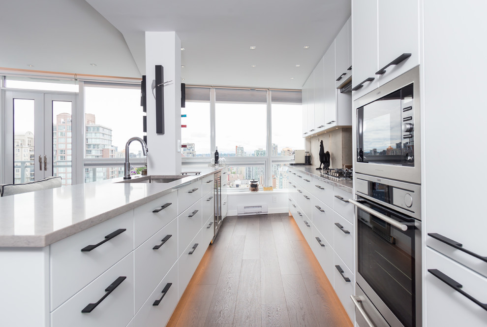This is an example of a mid-sized contemporary galley kitchen in Vancouver with flat-panel cabinets, white cabinets, light hardwood floors, an undermount sink, black appliances, with island, brown floor and grey benchtop.