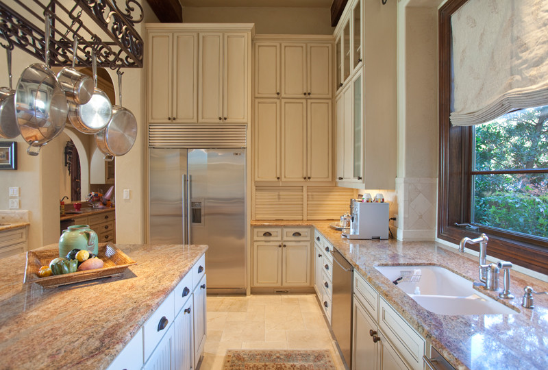 Photo of a large mediterranean u-shaped open plan kitchen in Austin with a drop-in sink, raised-panel cabinets, white cabinets, beige splashback, stone tile splashback, stainless steel appliances, limestone floors and with island.