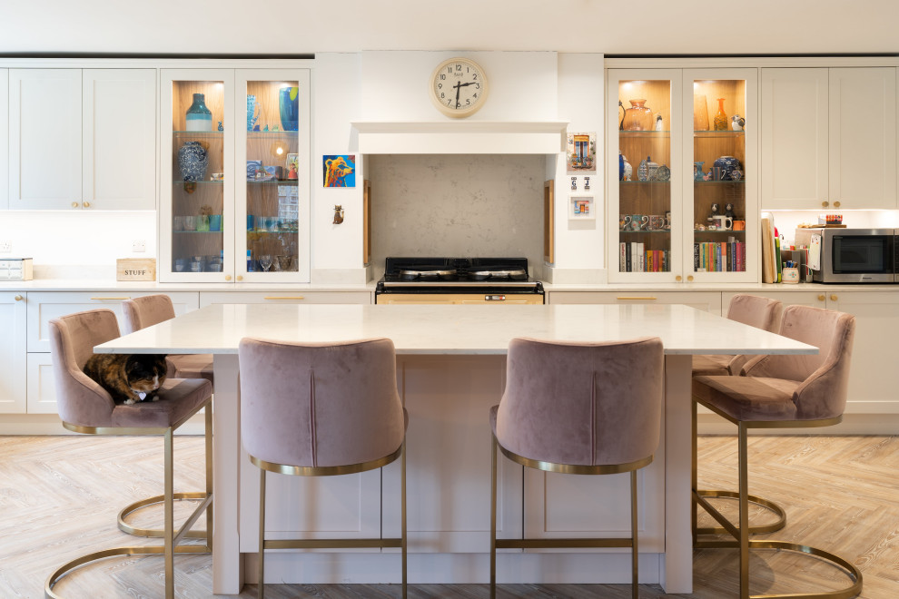 This is an example of a transitional galley kitchen in Hertfordshire with shaker cabinets, white cabinets, light hardwood floors, with island, beige floor and white benchtop.