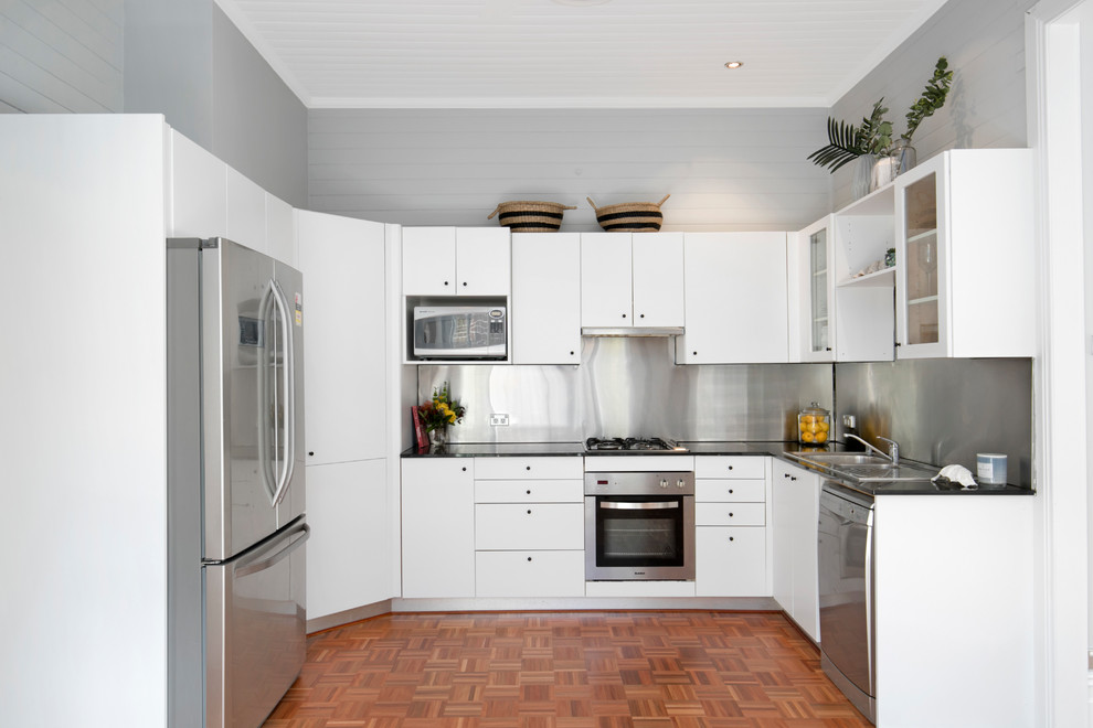 Photo of a modern u-shaped kitchen in Sydney with a double-bowl sink, stainless steel appliances, medium hardwood floors, flat-panel cabinets, white cabinets, granite benchtops, metal splashback, no island, brown floor and black benchtop.