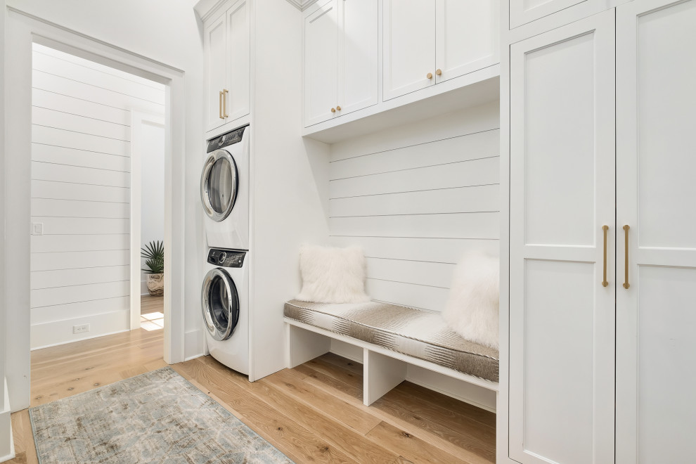 Design ideas for a large nautical single-wall utility room in Other with recessed-panel cabinets, white cabinets, medium hardwood flooring, a stacked washer and dryer and brown floors.