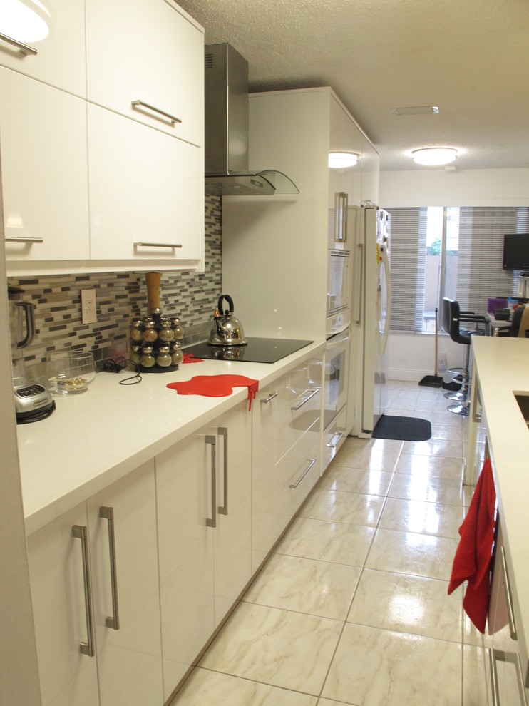 This is an example of a mid-sized modern galley eat-in kitchen in Miami with a double-bowl sink, flat-panel cabinets, white cabinets, quartz benchtops, multi-coloured splashback, glass sheet splashback, white appliances, porcelain floors and a peninsula.