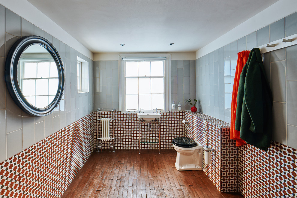 Photo of a mid-sized transitional 3/4 bathroom in London with a one-piece toilet, gray tile, red tile, white tile, medium hardwood floors, a pedestal sink, brown floor and white benchtops.