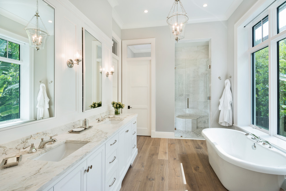 Transitional master bathroom in Other with recessed-panel cabinets, white cabinets, a freestanding tub, an alcove shower, white tile, grey walls, light hardwood floors, an undermount sink, brown floor, a hinged shower door and an enclosed toilet.
