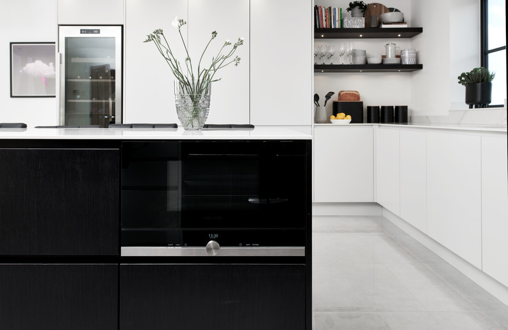 Photo of a modern open plan kitchen in Wiltshire with a single-bowl sink, flat-panel cabinets, dark wood cabinets, composite countertops, white splashback, glass sheet splashback, black appliances, porcelain flooring, an island, grey floors and white worktops.