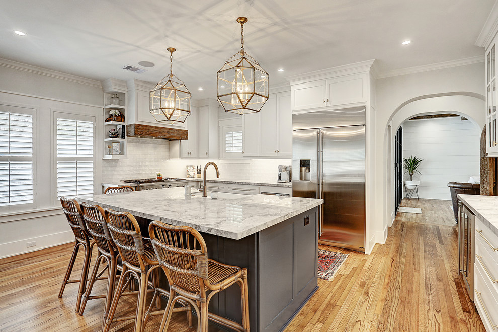 Inspiration for a country u-shaped kitchen in Houston with a farmhouse sink, shaker cabinets, white cabinets, white splashback, stainless steel appliances, medium hardwood floors, with island, brown floor and grey benchtop.