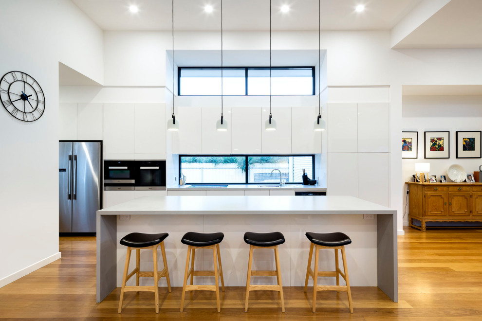 Inspiration for a large contemporary galley open plan kitchen in Adelaide with an undermount sink, flat-panel cabinets, white cabinets, window splashback, stainless steel appliances, with island, brown floor and grey benchtop.