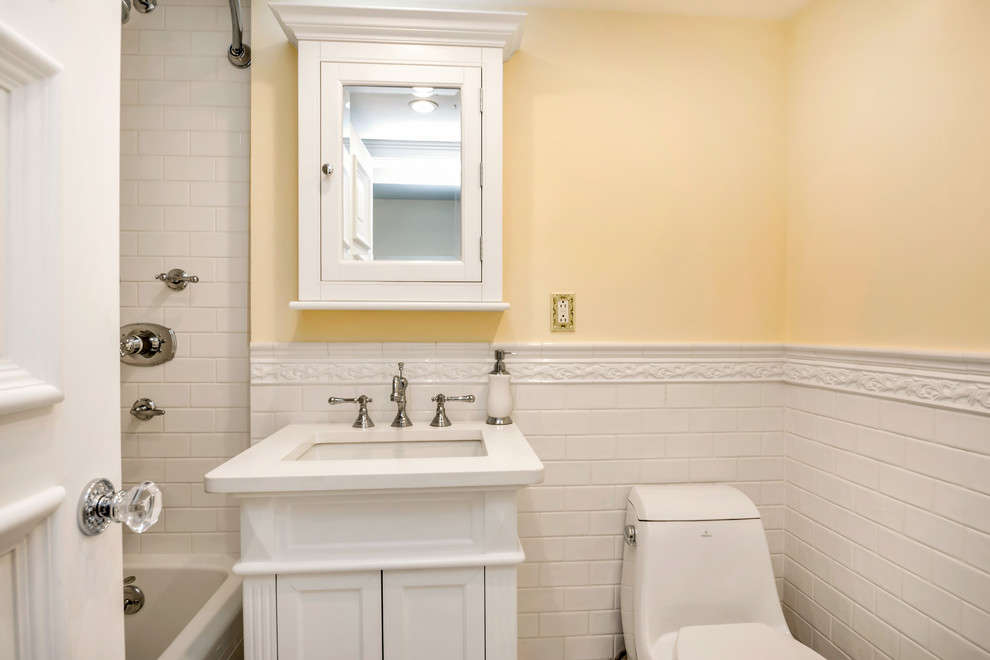 This is an example of a small traditional kids bathroom in New York with furniture-like cabinets, a one-piece toilet, white tile, stone tile, white cabinets, an alcove tub, a shower/bathtub combo, yellow walls, marble floors, an integrated sink and solid surface benchtops.