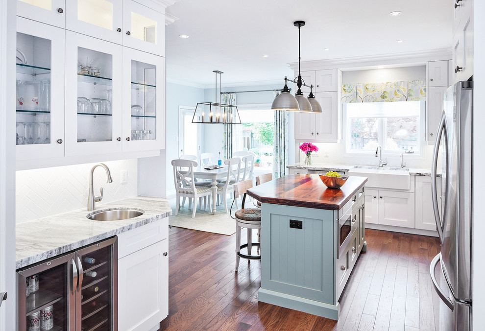 Example of a large transitional medium tone wood floor eat-in kitchen design in Toronto with a farmhouse sink, shaker cabinets, white cabinets, granite countertops, white backsplash, subway tile backsplash, stainless steel appliances and an island