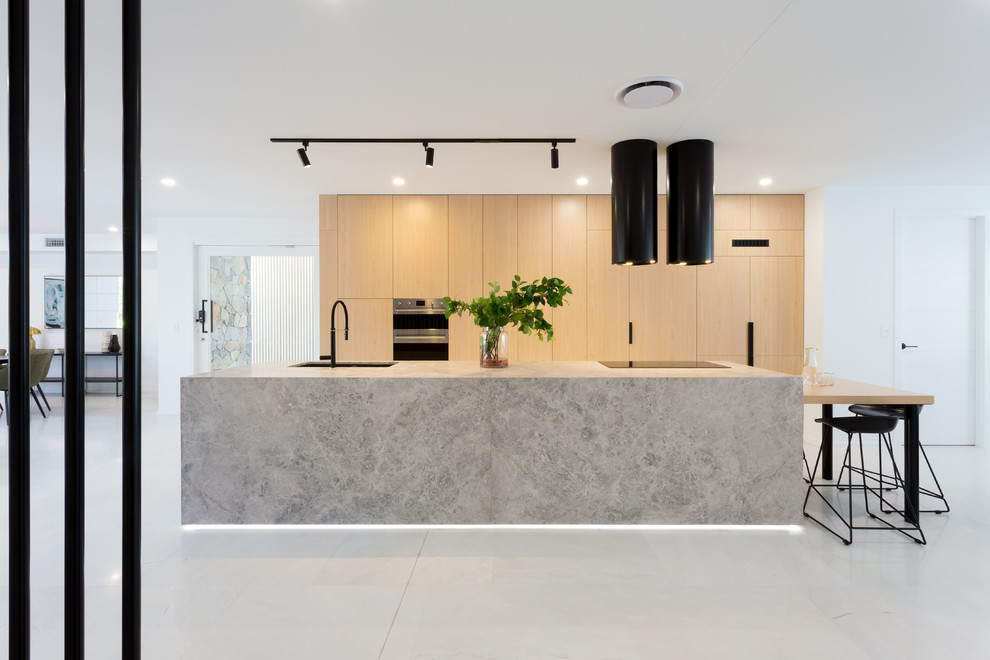 Large contemporary galley open plan kitchen in Brisbane with an undermount sink, light wood cabinets, limestone benchtops, black appliances, marble floors, with island, white floor, grey benchtop and flat-panel cabinets.